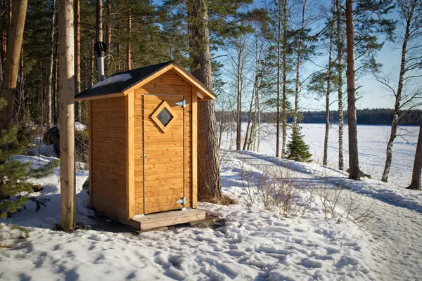An outhouse made of yellow pine with a diamond shaped window cut into the door sits amongst trees in the snowy woods.